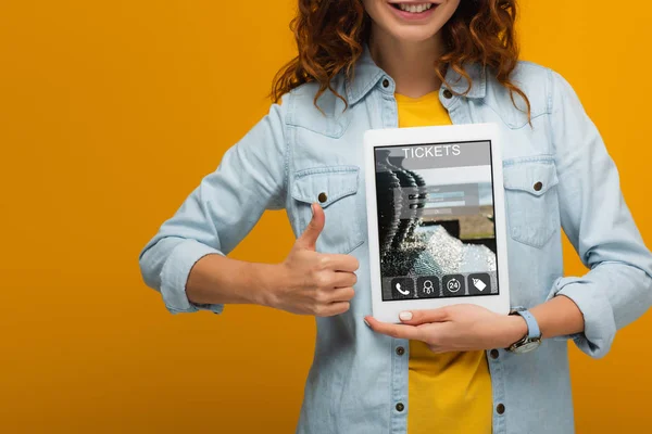 Обрізаний вид веселої кучерявої жінки, що тримає цифровий планшет з додатком для квитків на екрані і показує великий палець вгору ізольовано на помаранчевому — стокове фото