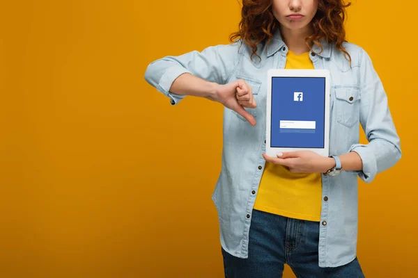 Vue recadrée de femme bouclée bouleversée tenant tablette numérique avec application facebook à l'écran et montrant pouce vers le bas sur orange — Photo de stock