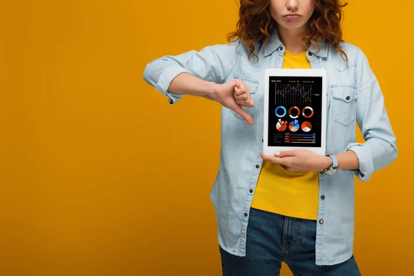 Cropped view of upset curly woman holding digital tablet with charts and graphs on screen and showing thumb down on orange — Stock Photo