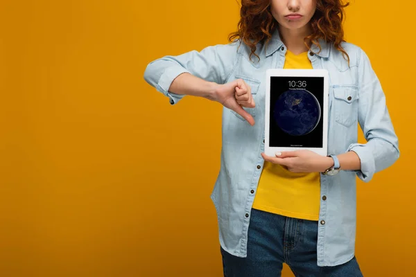 Cropped view of upset curly woman holding digital tablet with lock screen and showing thumb down on orange — Stock Photo