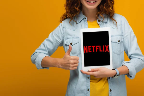 Обрезанный вид веселой кудрявой женщины, держащей цифровой планшет с приложением netflix на экране и показывающей большой палец, изолированный на оранжевом — стоковое фото