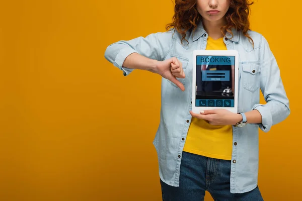 Ausgeschnittene Ansicht einer aufgebrachten lockigen Frau, die ein digitales Tablet mit Buchungs-App auf dem Bildschirm hält und den Daumen nach unten auf orange zeigt — Stockfoto