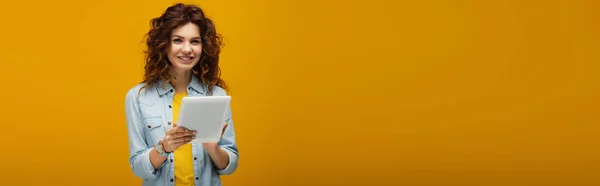 Panoramaaufnahme eines fröhlichen lockigen rothaarigen Mädchens, das in die Kamera blickt und ein digitales Tablet auf orange hält — Stockfoto