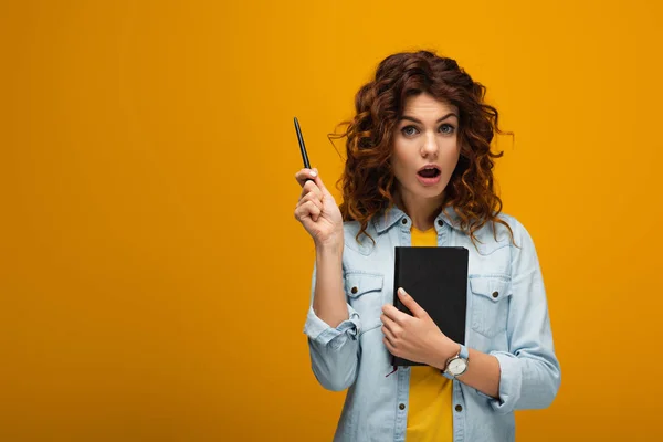 Chica pelirroja sorprendida sosteniendo portátil y pluma y mirando a la cámara en naranja - foto de stock