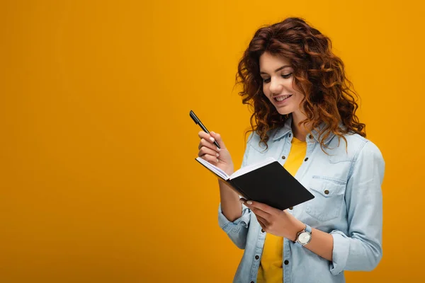 Cheerful redhead young woman holding notebook and pen on orange — Stock Photo