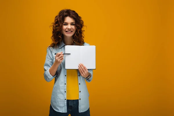 Smiling redhead young woman pointing at blank notebook with pen on orange — Stock Photo
