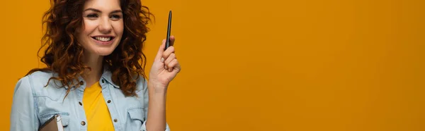 Colpo panoramico di rossa sorridente giovane donna in possesso di notebook e penna su arancione — Foto stock