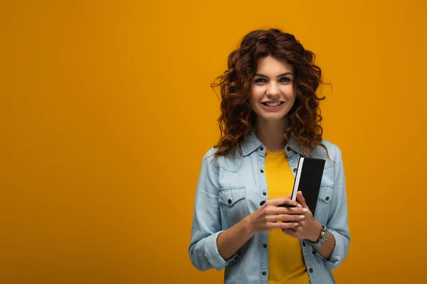 Cheerful and beautiful redhead young woman holding notebook and pen on orange — Stock Photo