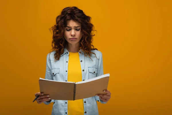 Upset redhead young woman looking at notebook and holding pen on orange — Stock Photo