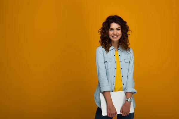 Happy redhead girl holding laptop and smiling on orange — Stock Photo