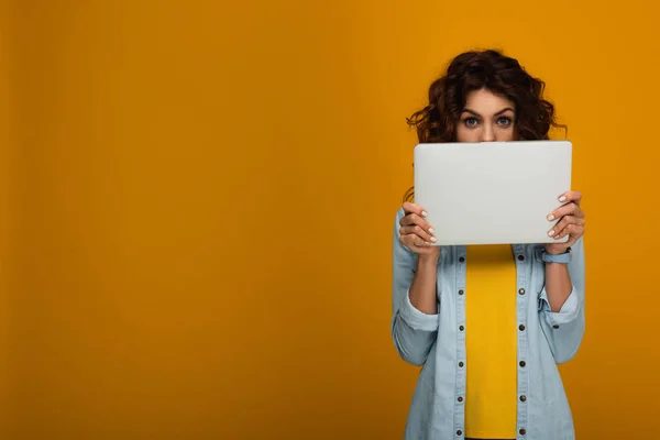 Curly redhead girl covering face with laptop on orange — Stock Photo