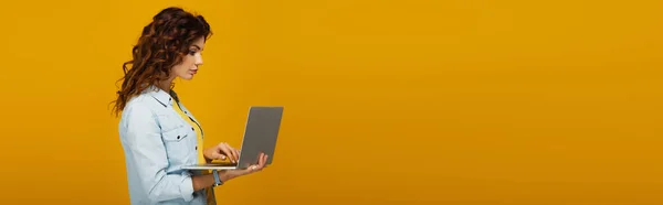 Plano panorámico de hermosa y rizada chica pelirroja utilizando el ordenador portátil aislado en naranja - foto de stock