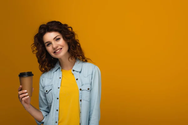 Happy redhead girl holding paper cup with drink isolated on orange — Stock Photo