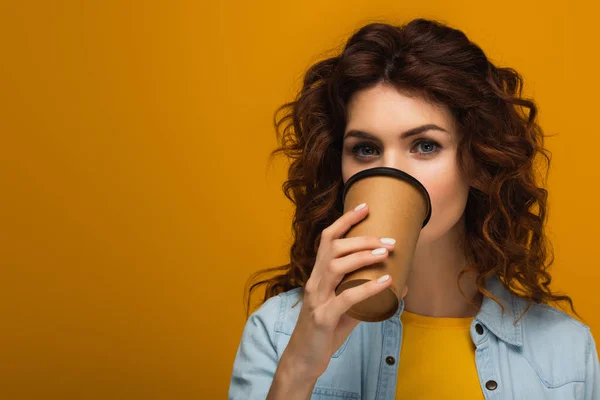 Curly redhead girl covering face with paper cup while drinking on orange — Stock Photo