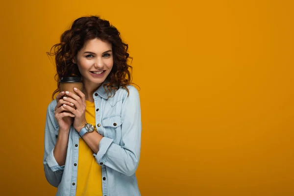 Happy curly redhead girl holding paper cup while smiling on orange — Stock Photo