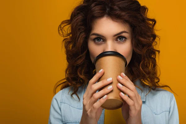 Pelirroja rizada cubriendo la cara con taza de papel mientras bebe aislado en naranja - foto de stock