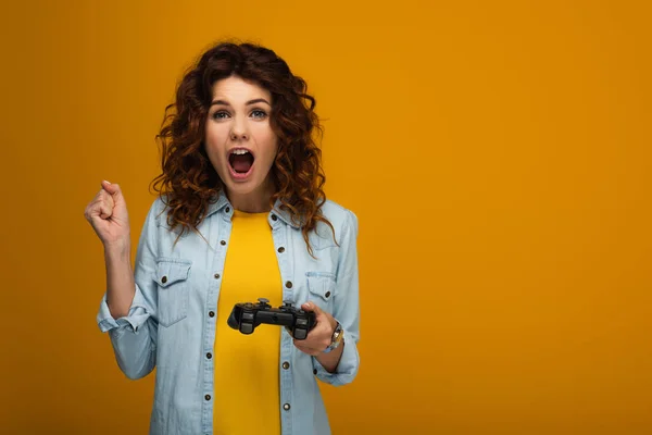 Emotional curly woman gesturing and screaming while holding joystick on orange — Stock Photo