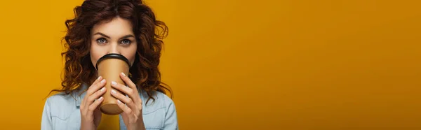 Panoramic shot of curly redhead girl covering face with paper cup while drinking on orange — Stock Photo