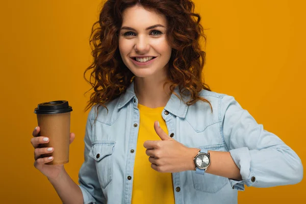 Heureux bouclé rousse fille tenant café pour aller et montrant pouce isolé sur orange — Photo de stock