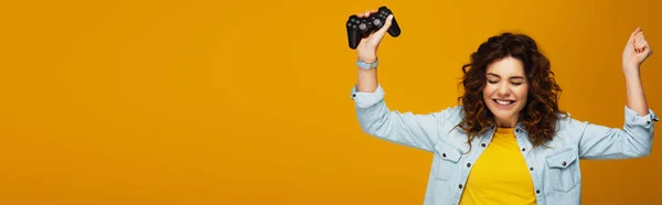 Plano panorámico de alegre chica pelirroja rizada gesto mientras sostiene el joystick en naranja - foto de stock