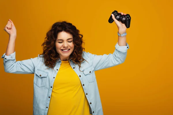 Cheerful curly redhead girl gesturing while holding joystick on orange — Stock Photo