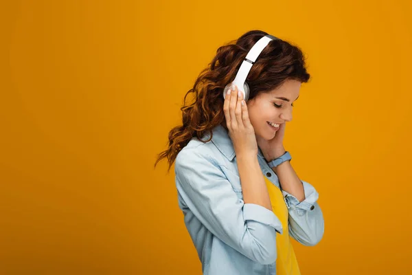 Chica pelirroja feliz sonriendo mientras toca los auriculares en naranja - foto de stock