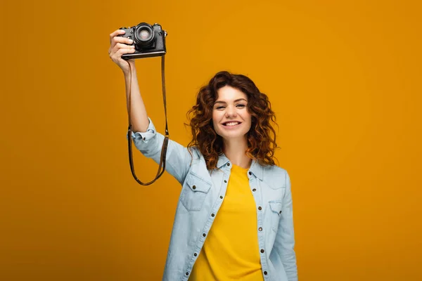 Joyeux photographe rousse tenant appareil photo numérique au-dessus de la tête sur orange — Photo de stock