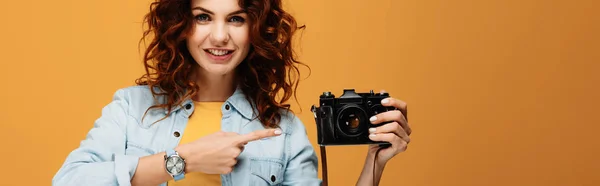 Fotografía panorámica de fotógrafo pelirrojo alegre apuntando con el dedo a la cámara digital en naranja - foto de stock