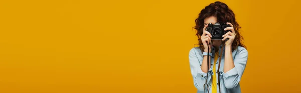 Панорамний знімок кучерявого рудого фотографа, що закриває обличчя цифровою камерою ізольовано на помаранчевому — стокове фото