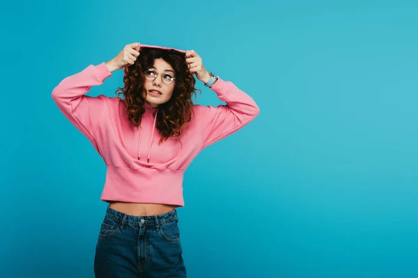 Attractive redhead girl wearing hood while standing in pink sweatshirt on blue — Stock Photo