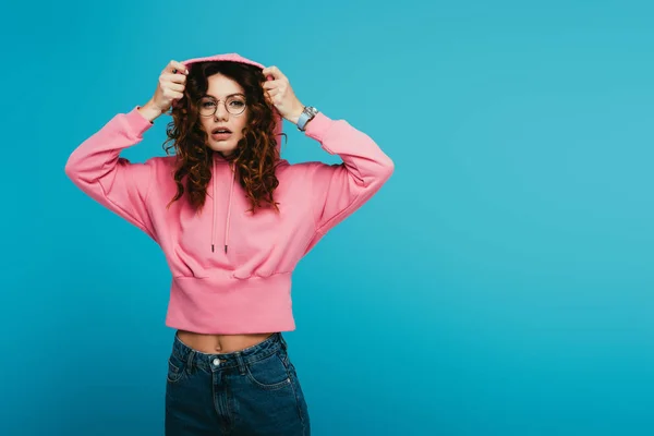 Surprised redhead girl wearing hood while standing in pink sweatshirt on blue — Stock Photo