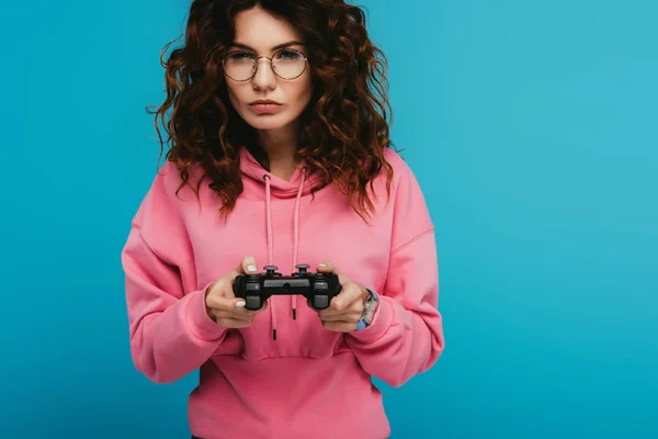 Серйозна кучерява руда дівчина грає в відеогру, тримаючи джойстик на синьому — стокове фото