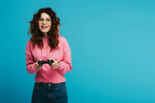 Feliz encaracolado ruiva menina jogar vídeo game enquanto segurando joystick no azul — Fotografia de Stock