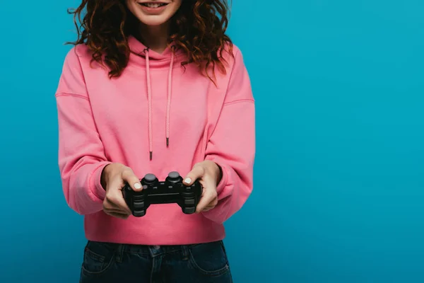 Vista cortada de menina ruiva encaracolado alegre jogando videogame enquanto segurando joystick no azul — Fotografia de Stock