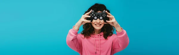 Panoramic shot of happy curly redhead girl covering face with joystick on blue — Stock Photo