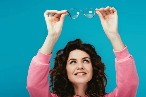 Веселая кудрявая девушка, смотрящая на очки, изолированные на голубом — стоковое фото