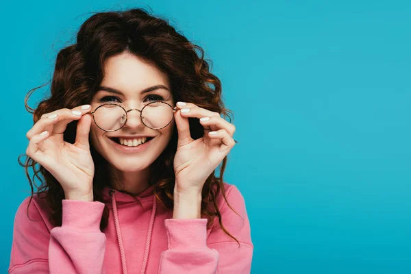 Cheerful curly redhead girl touching glasses isolated on blue — Stock Photo