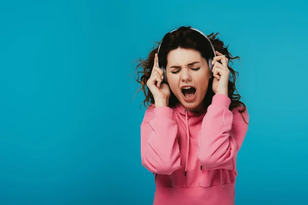 Mulher encaracolado atraente cantando enquanto ouve música em fones de ouvido em azul — Fotografia de Stock
