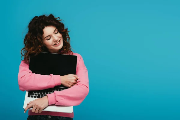 Alegre chica pelirroja rizada abrazando portátil con pantalla en blanco, mientras que de pie en azul - foto de stock