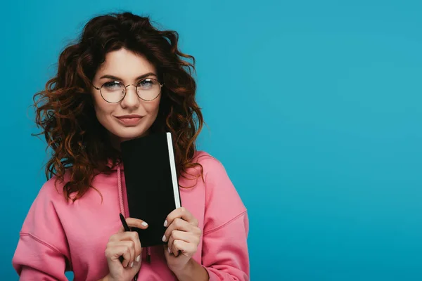 Alegre chica rizada en gafas sosteniendo portátil y pluma aislado en azul - foto de stock