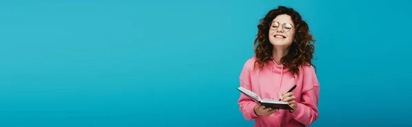 Panoramic shot of happy curly girl in glasses holding notebook and pen isolated on blue — Stock Photo