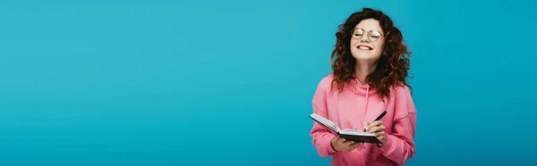 Tiro panorâmico de menina encaracolado feliz em óculos segurando notebook e caneta isolada em azul — Fotografia de Stock