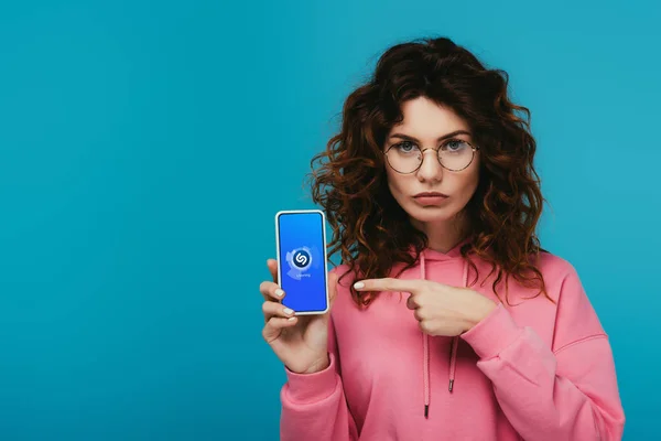 Attrayant rousse bouclée fille pointant du doigt à smartphone avec application shazam sur l'écran isolé sur bleu — Photo de stock