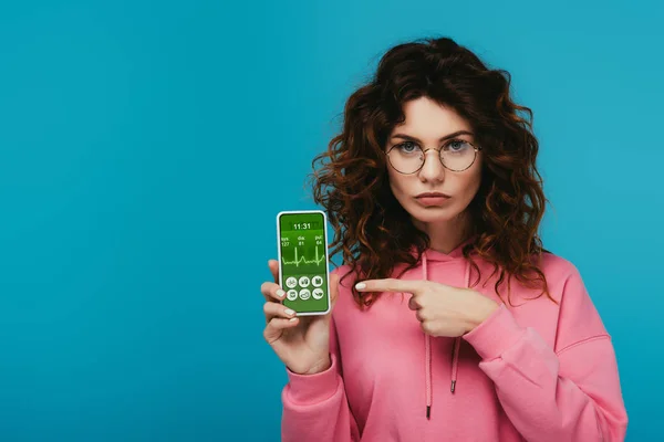Atractiva chica pelirroja rizada apuntando con el dedo en el teléfono inteligente con aplicación de salud electrónica en la pantalla aislada en azul - foto de stock