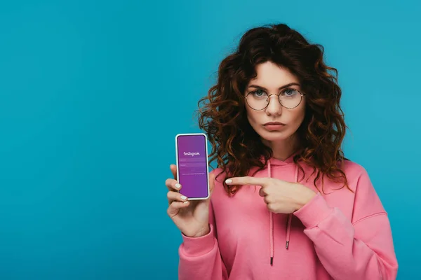 Atractiva chica pelirroja rizada apuntando con el dedo en el teléfono inteligente con aplicación instagram en la pantalla aislada en azul - foto de stock