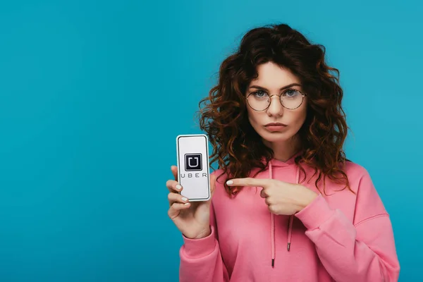 Atractiva chica pelirroja rizada apuntando con el dedo en el teléfono inteligente con aplicación uber en la pantalla aislada en azul - foto de stock