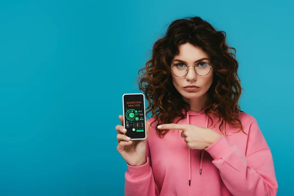 Привлекательная кудрявая рыжая девушка указывая пальцем на смартфон с маркетинговым анализом на экране изолированы на синий — Stock Photo