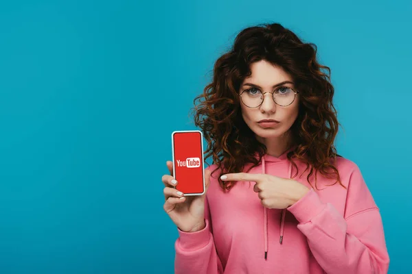 Attraente ragazza rossa riccia che punta con il dito allo smartphone con l'applicazione youtube sullo schermo isolato su blu — Foto stock