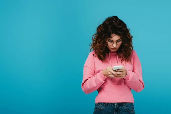 Jolie fille bouclée dans des lunettes en utilisant smartphone sur bleu — Photo de stock