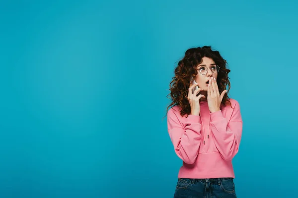 Surprised curly girl in glasses covering mouth while talking on smartphone on blue — Stock Photo